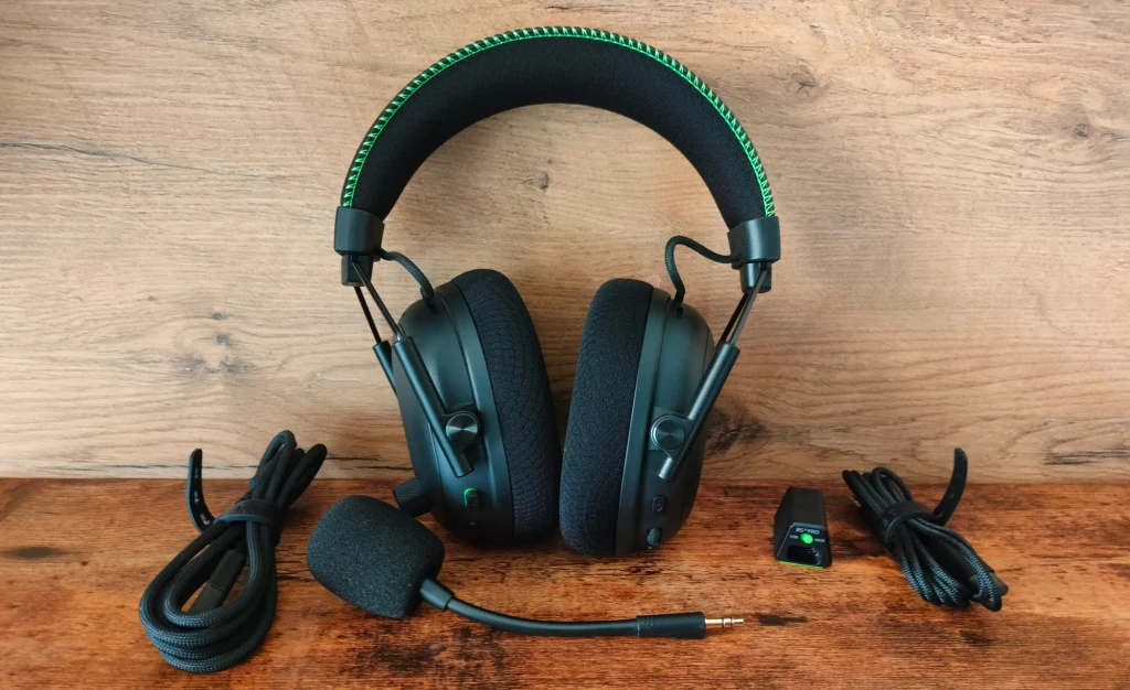 Razer Blackshark V3 Pro Czarne, nauszne słuchawki gamingowe z grubym pałąkiem ozdobionym zielonym przeszyciem, mikrofon na wysięgniku, dwa przewody oraz odbiornik USB ułożone na drewnianym blacie.