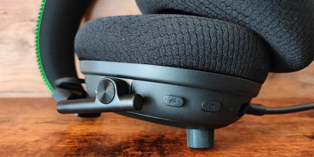 Razer Blackshark V3 Pro Duży, miękki nausznik słuchawek gamingowych z widocznym mikrofonem, przyciskiem wyciszenia dźwięku i pokrętłem regulacji głośności, na tle drewnianej powierzchni.