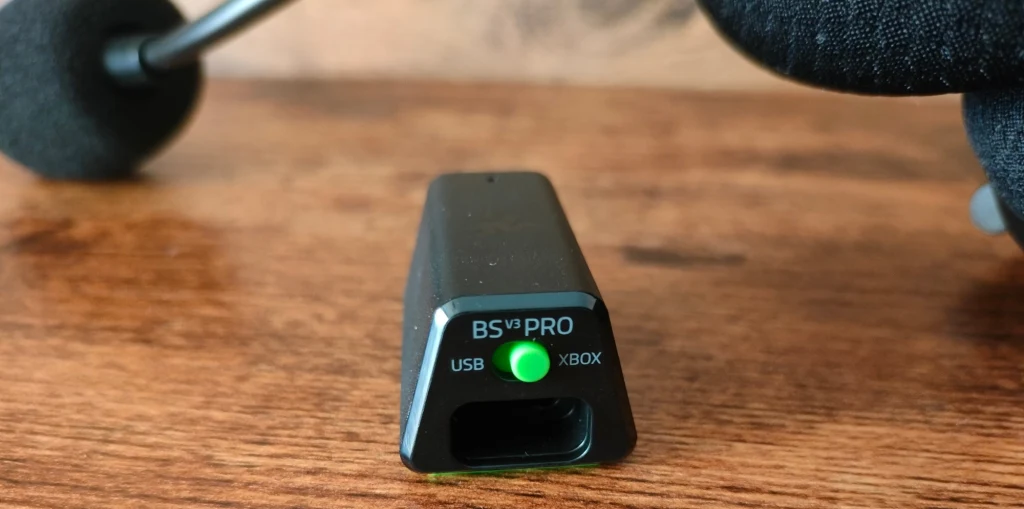 Razer Blackshark V3 Pro Adapter BS PRO z podłączeniem USB i wskaźnikiem diody, leży na drewnianej powierzchni. W tle widoczne części zestawu słuchawkowego, w tym mikrofon i nausznik.