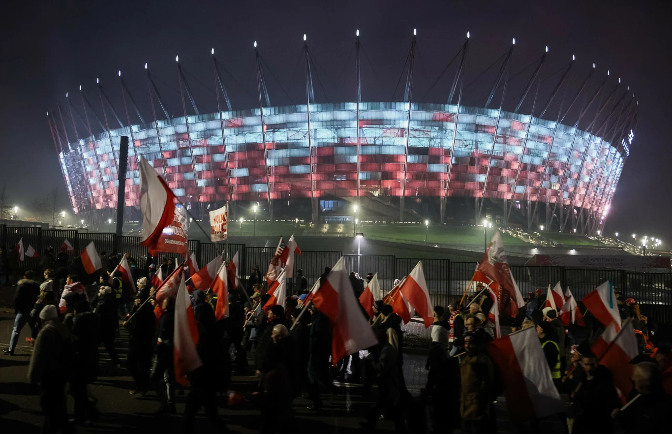 Tłum ludzi niosących polskie flagi maszeruje przed nowocześnie podświetlonym Stadionem Narodowym w Warszawie, na którym światła tworzą biało-czerwony wzór nawiązujący do barw narodowych Polski.