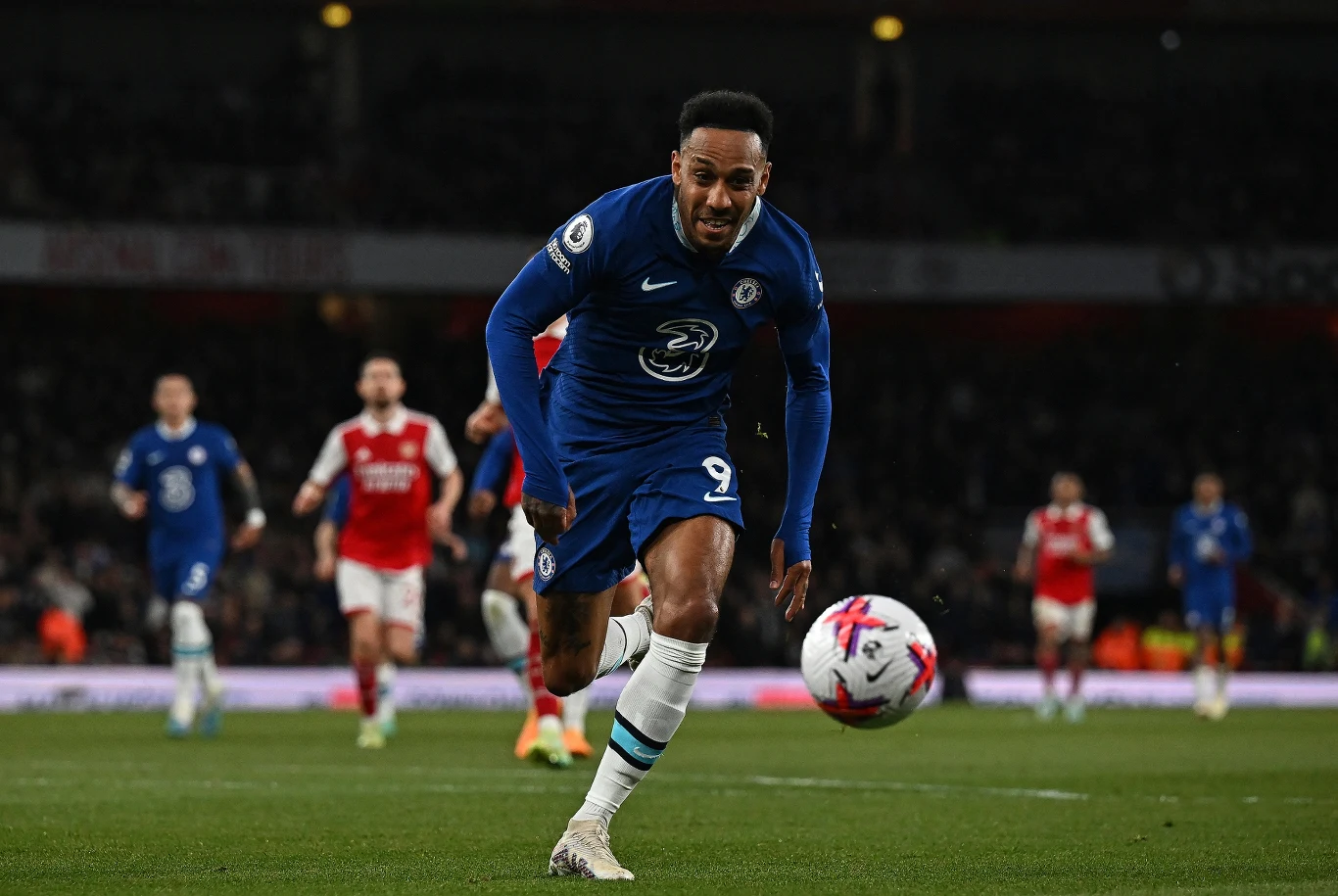 Piłkarz w niebieskiej koszulce Chelsea biegnie za piłką po murawie stadionu podczas meczu piłkarskiego, w tle widoczni zawodnicy drużyny przeciwnej ubrani w czerwono-białe stroje oraz kibice na trybunach.