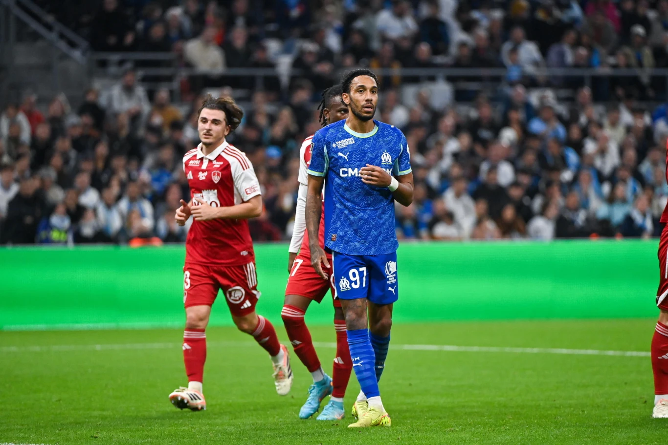 Piłkarz w niebieskiej koszulce drużyny Olympique Marsylia idzie po murawie obok zawodników drużyny przeciwnej ubranych na czerwono, w tle widoczna trybuna pełna kibiców i jasnozielona reklama świetlna przy bocznej linii.