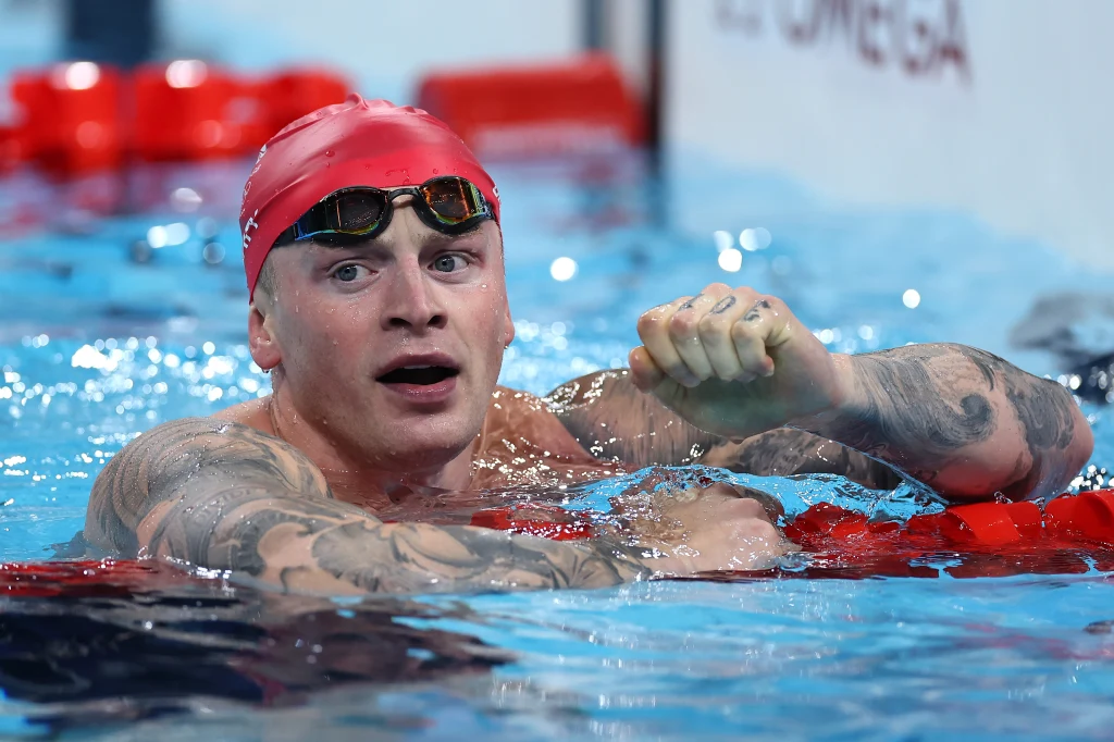 Adam Peaty Zawodowy pływak w czepku i okularach pływackich wynurza się z basenu, unosząc zaciśniętą pięść w geście triumfu, widoczne tatuaże na ramionach i intensywna mimika twarzy wskazują na sportowe emocje.