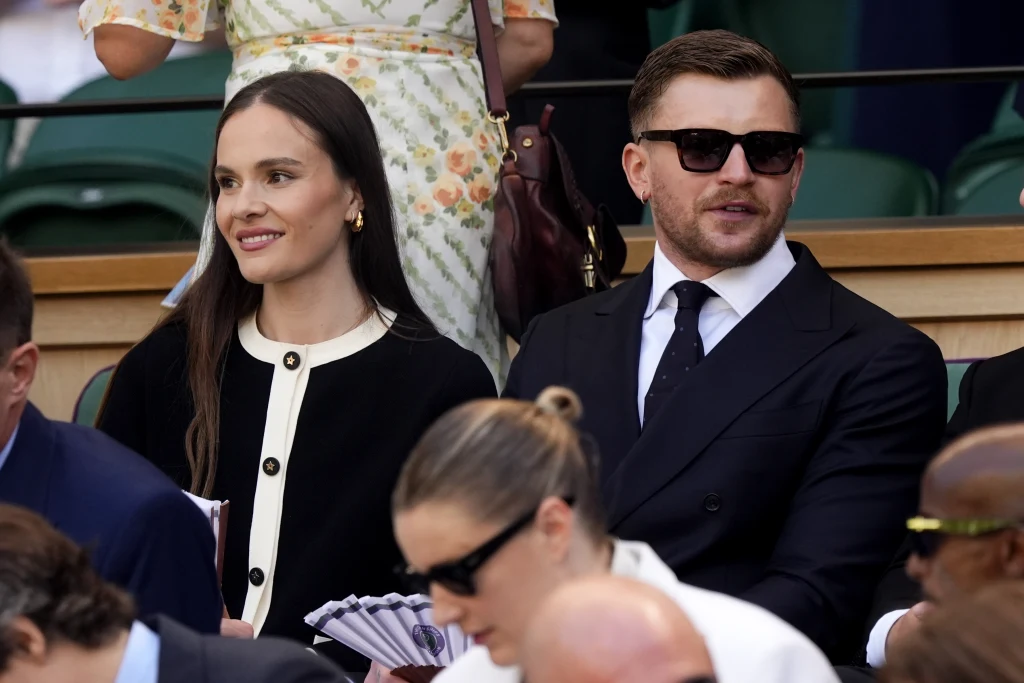 Holly Ramsay i Adam Peaty Elegancka kobieta z długimi, brązowymi włosami i uśmiechem siedzi obok mężczyzny w ciemnych okularach oraz garniturze, w tle widać innych uczestników wydarzenia sportowego.
