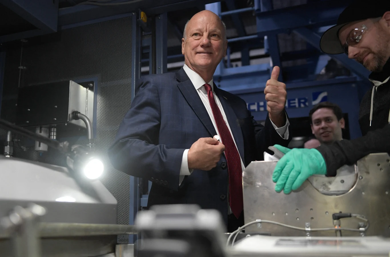 Mężczyzna w garniturze uśmiecha się i pokazuje gest kciuka w górę podczas wizyty w zakładzie przemysłowym, obok niego pracownicy w strojach roboczych, w tle widoczne są elementy techniczne instalacji.