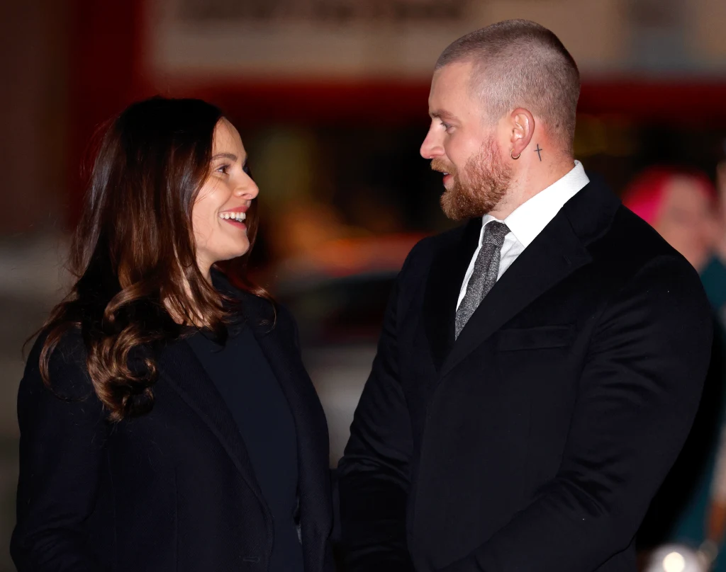 Holly Ramsay i Adam Peaty Para ubrana w eleganckie, ciemne stroje stoi twarzą w twarz, uśmiechając się do siebie podczas wieczornego wydarzenia, w tle rozmyte światła miasta.