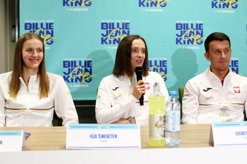 Trzy osoby ubrane w sportowe stroje z polskimi emblematami siedzą przy stole podczas konferencji prasowej, środkowa postać trzyma mikrofon, w tle baner Billie Jean King Cup.