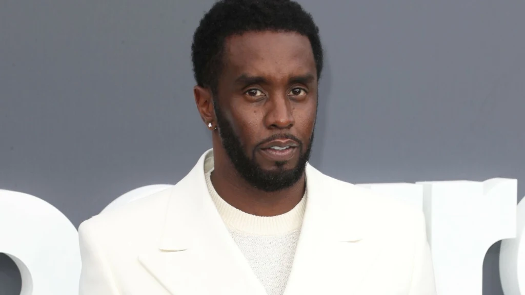 Sean "Diddy" Combs Mężczyzna w białym garniturze i kremowym swetrze stoi na tle szarej ściany z białymi elementami. Ma krótką, czarną fryzurę i krótko przystrzyżoną brodę. Spogląda poważnie w stronę obiektywu, nosi kolczyk w lewym uchu.