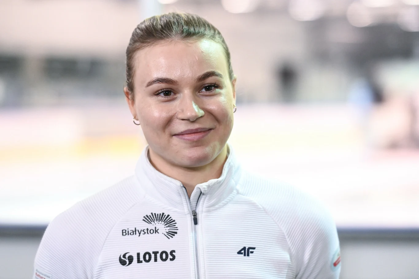 Natalia Maliszewska Uśmiechnięta młoda kobieta w sportowej białej bluzie z logotypami Białystok, Lotos i 4F, tło rozmyte, neutralne barwy.