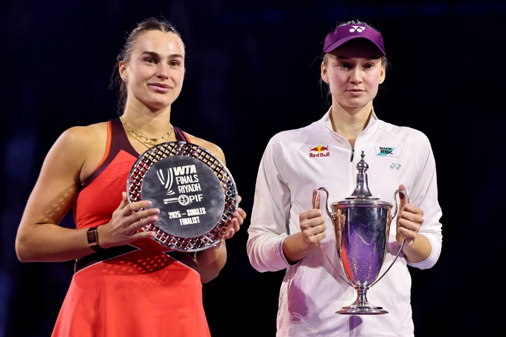 Aryna Sabalenka i Jelena Rybakina Dwie kobiety trzymające trofea, jedna w sportowej czerwonej sukience z talerzem za drugie miejsce WTA Finals, druga w białej sportowej koszulce i czapce z pucharem za pierwsze miejsce.