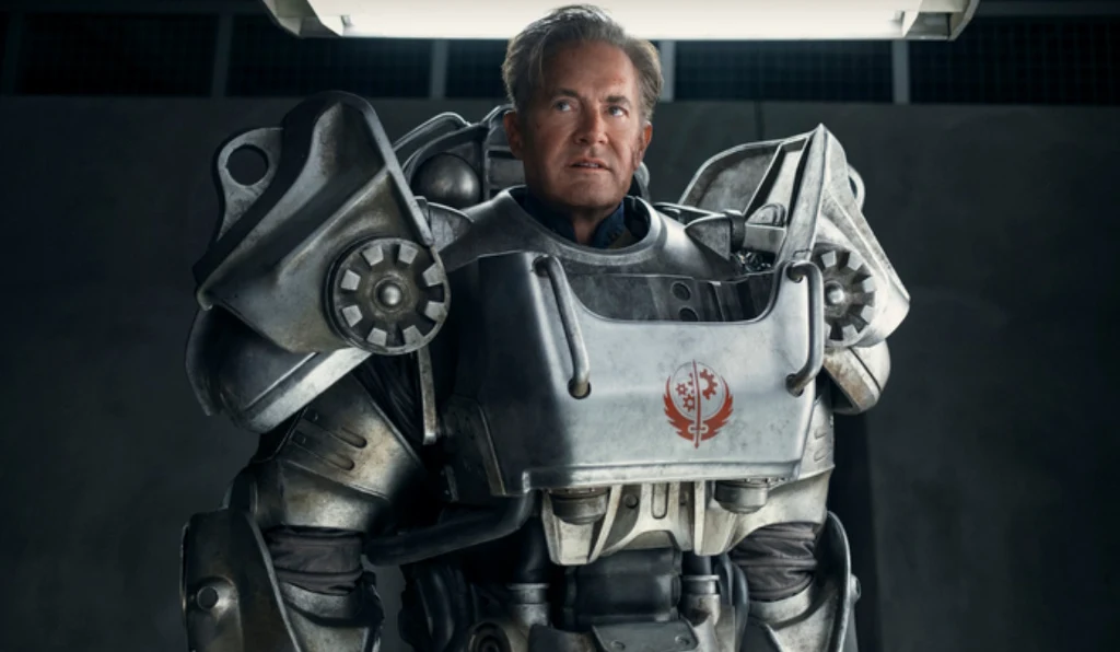 Mężczyzna ubrany w ciężki, futurystyczny pancerz bojowy stoi pod jasnym światłem na neutralnym tle. Zbroja jest masywna, metalowa, z widocznym emblematem na piersi oraz licznymi detalami technicznymi.