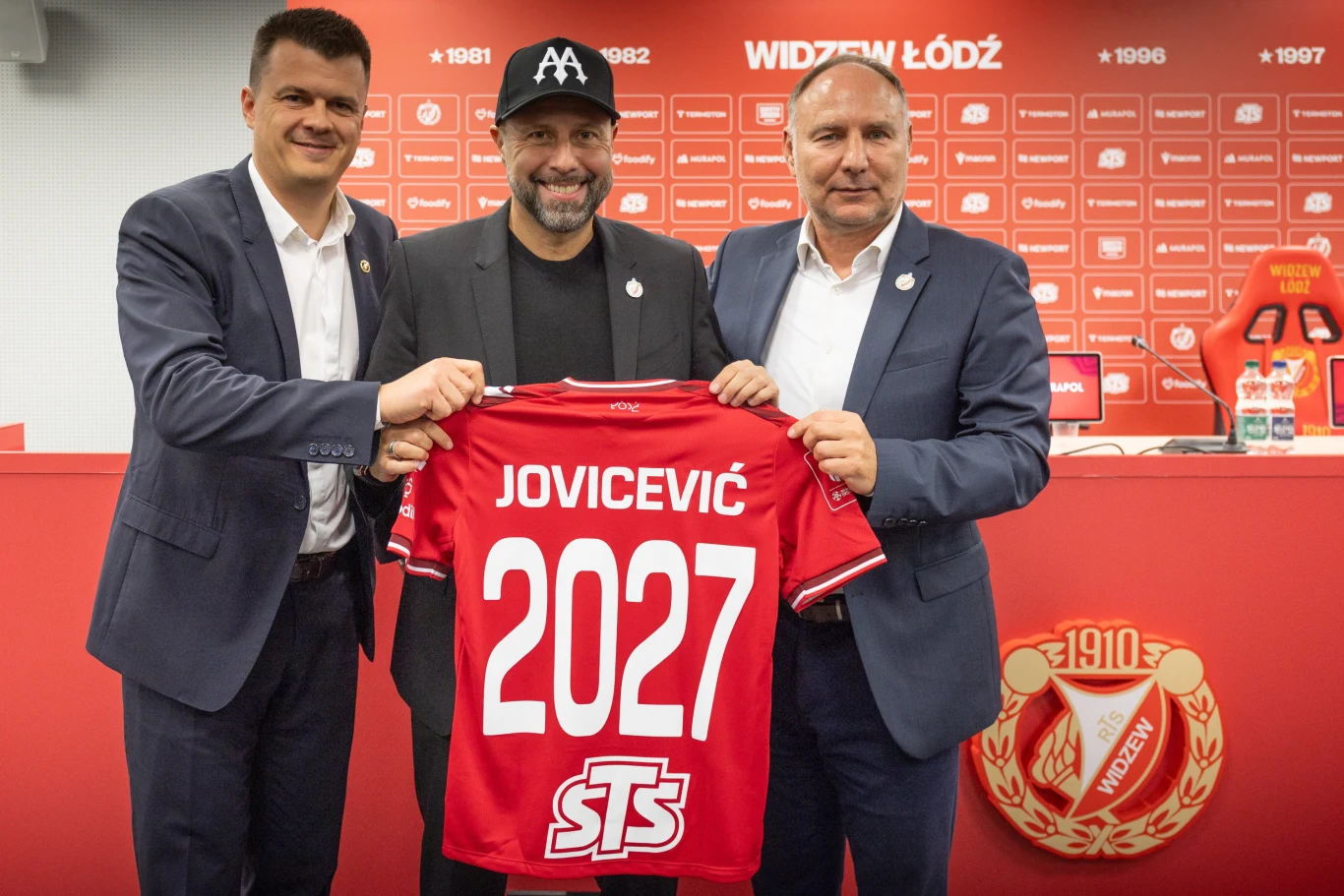 Od lewej: Mindaugas Nikolicius, Igor Jovićević, Dariusz Adamczuk Trzech mężczyzn w garniturach stoi obok siebie w jasnym pomieszczeniu klubowym. Dwóch z nich trzyma czerwoną koszulkę piłkarską z napisem 'JOVIĆEVIĆ 2027' i logiem sponsora STS. W tle widoczna czerwona ścianka z logotypami oraz duży herb klubu Widzew Ł...