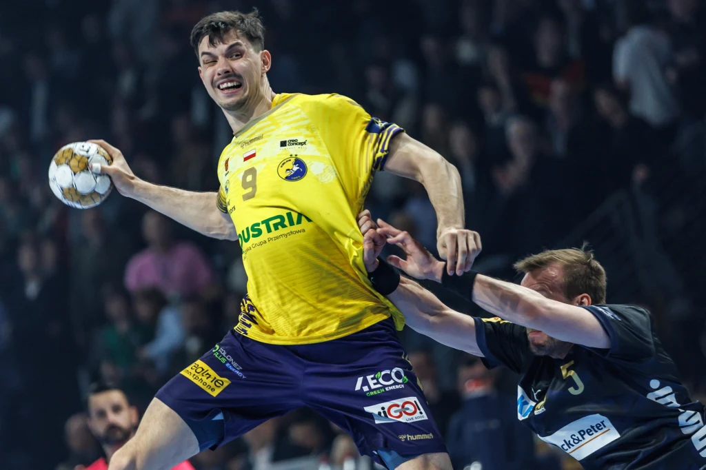 Szymon Sićko Dwóch zawodników podczas dynamicznej akcji piłki ręcznej, jeden z nich w żółtej koszulce unosi się w powietrzu z piłką, broniąc się przed przeciwnikiem ubranego w ciemny strój, wokół zamazana publiczność na trybunach.