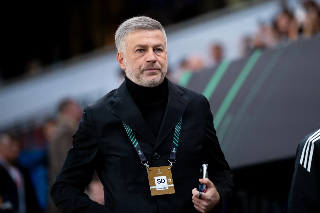 Mężczyzna z siwą brodą ubrany w czarny golf i marynarkę, z identyfikatorem na szyi, stoi na tle rozmytego stadionu sportowego, trzymając notes i długopis w dłoni.