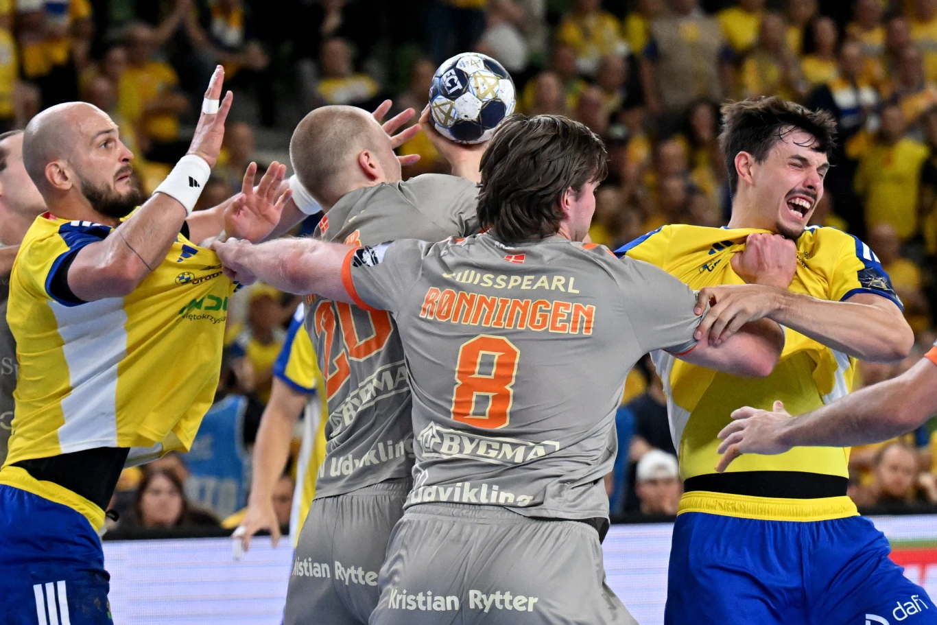 Zacięta walka zawodników podczas meczu piłki ręcznej. Gracze w żółto-niebieskich i szaro-pomarańczowych strojach próbują przejąć piłkę, w tle widownia.