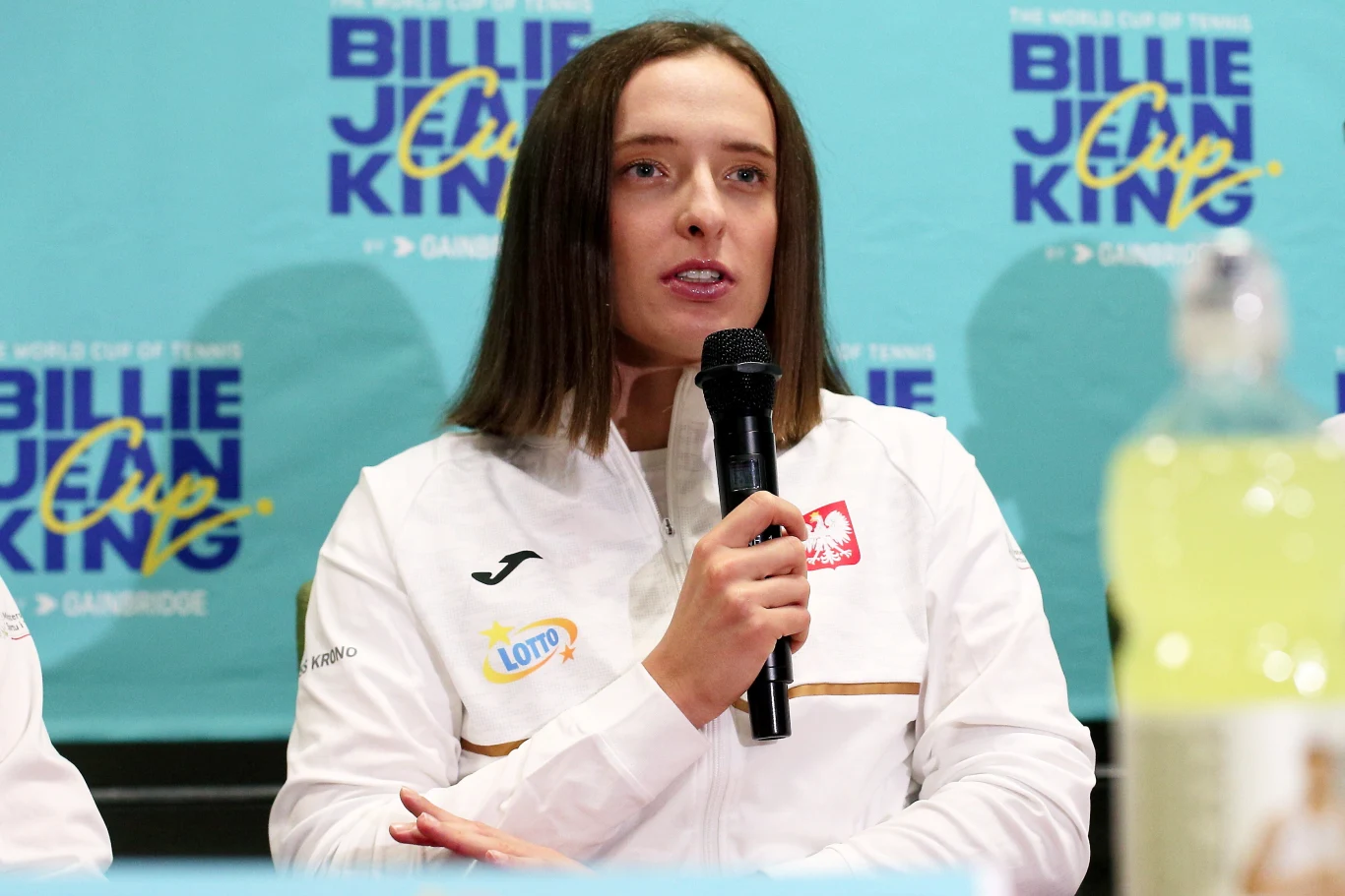 Kobieta w sportowym stroju z logo Lotto i polską flagą trzyma mikrofon podczas konferencji prasowej na tle niebieskiego banera z napisem Billie Jean King Cup.