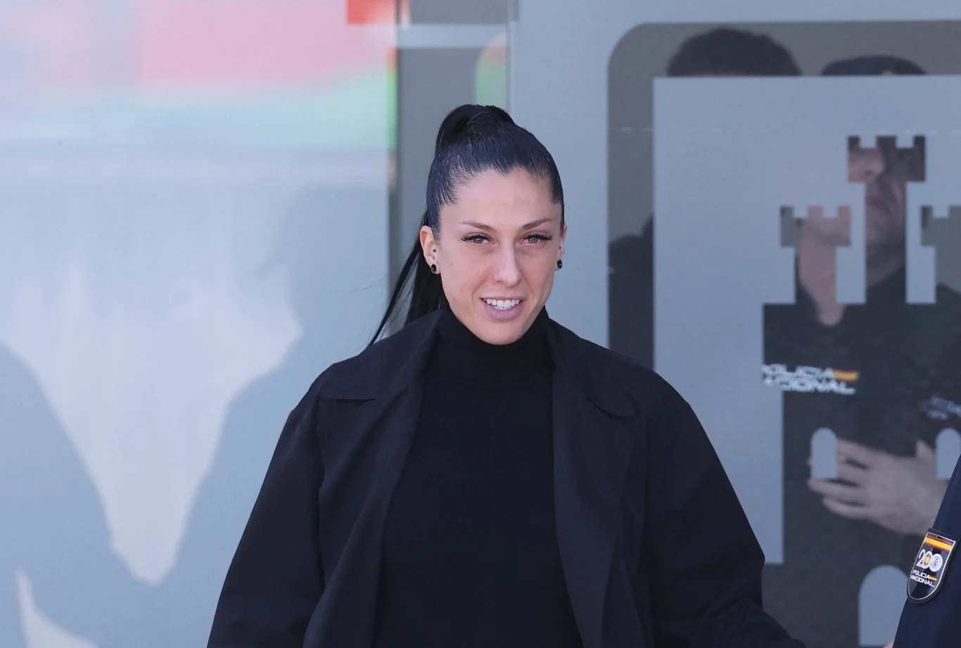 Kobieta z długimi, ciemnymi włosami spiętymi w kucyk, ubrana na czarno, stoi na tle szarej ściany z geometrycznymi wzorami oraz logotypem.