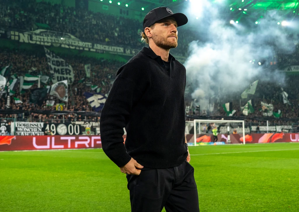 Mężczyzna w czarnej czapce i ciemnych ubraniach stoi na zielonej murawie stadionu, w tle widać rozświetlone trybuny pełne kibiców, flagi i dym, panuje intensywna atmosfera wydarzenia sportowego.