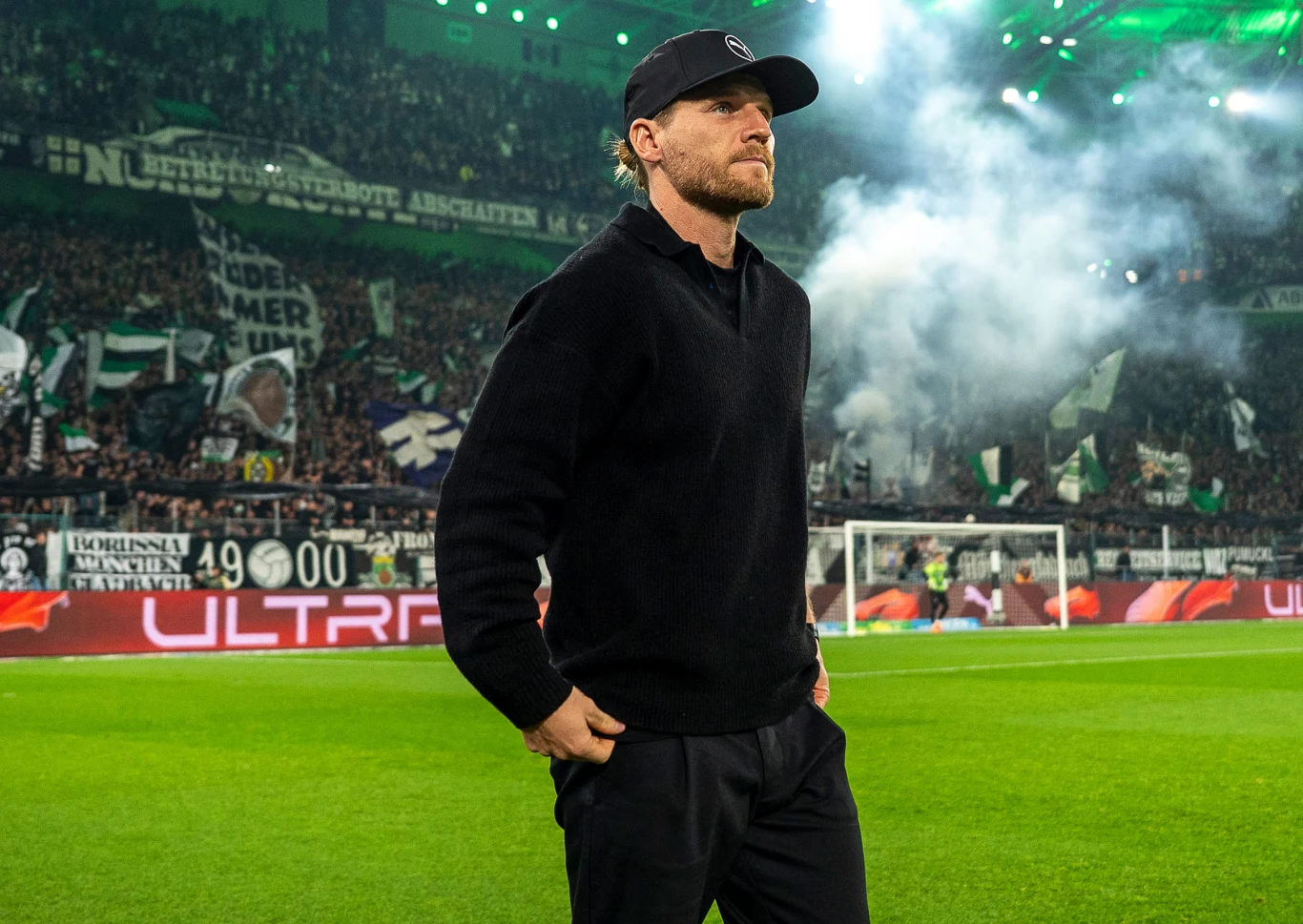 Mężczyzna w czarnej czapce i ciemnych ubraniach stoi na zielonej murawie stadionu, w tle widać rozświetlone trybuny pełne kibiców, flagi i dym, panuje intensywna atmosfera wydarzenia sportowego.