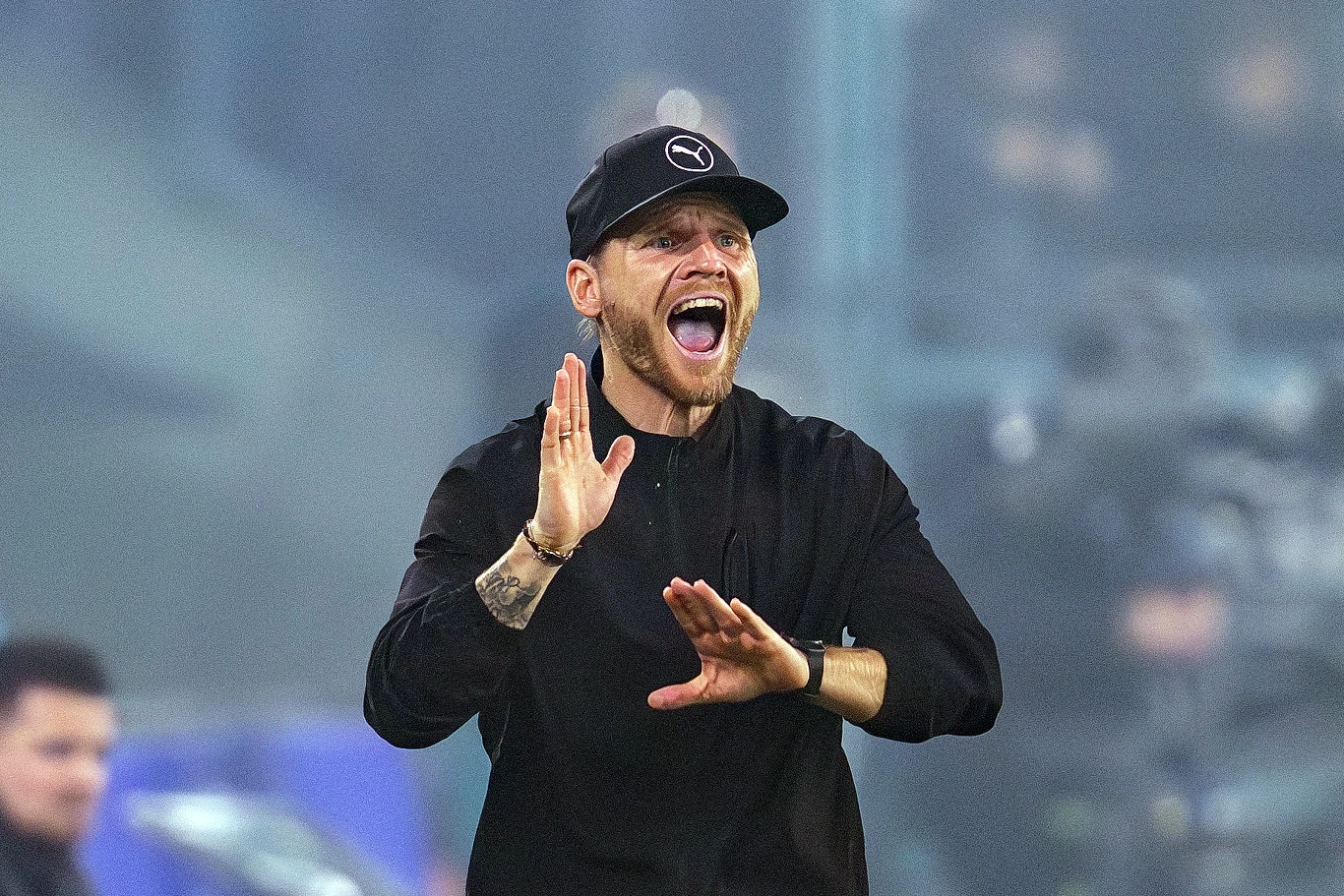 Eugen Polanski Mężczyzna w czarnej czapce i bluzie, z szeroko otwartymi ustami, wykonujący gest rękoma jakby instruował lub motywował kogoś, rozgrywająca się dynamiczna scena na tle nastrojowego oświetlenia.