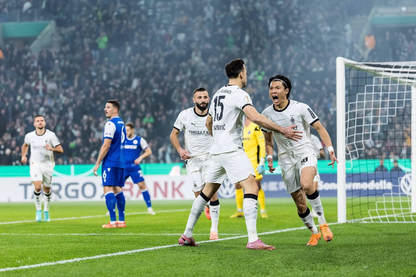 Piłkarze Borussii Monchengladbach Dwaj piłkarze świętują zdobycie bramki na tle tłumu kibiców i innych zawodników na boisku, jeden z nich wyraża silne emocje zwycięstwa. W tle widoczna bramka oraz rywale ubrane w niebieskie stroje.