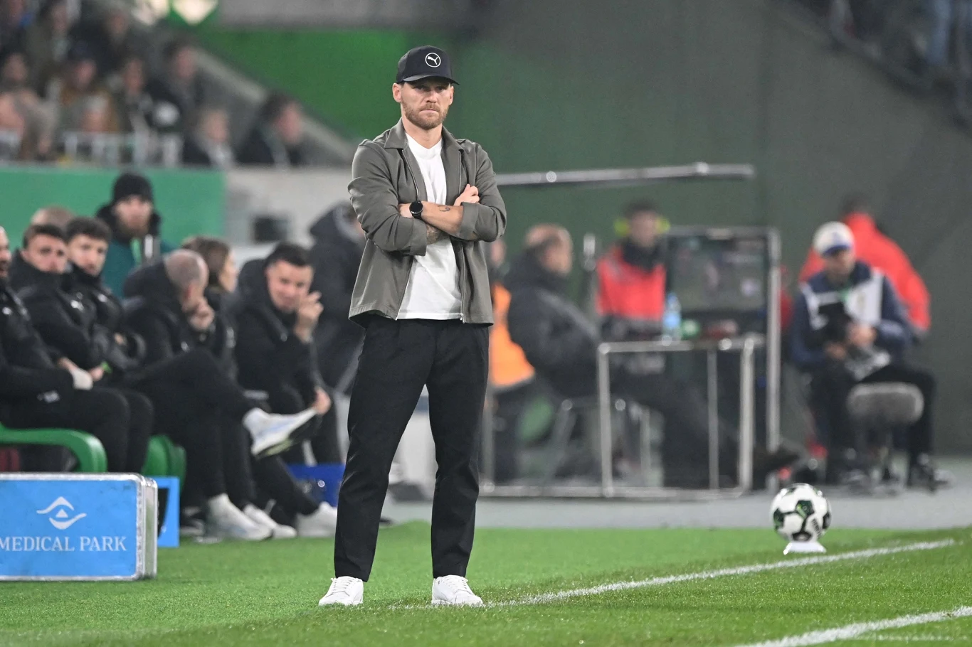 Mężczyzna w sportowym ubraniu i czapce z daszkiem stoi z założonymi rękami przy linii bocznej boiska piłkarskiego, w tle widoczni siedzący członkowie sztabu trenerskiego oraz elementy stadionu.