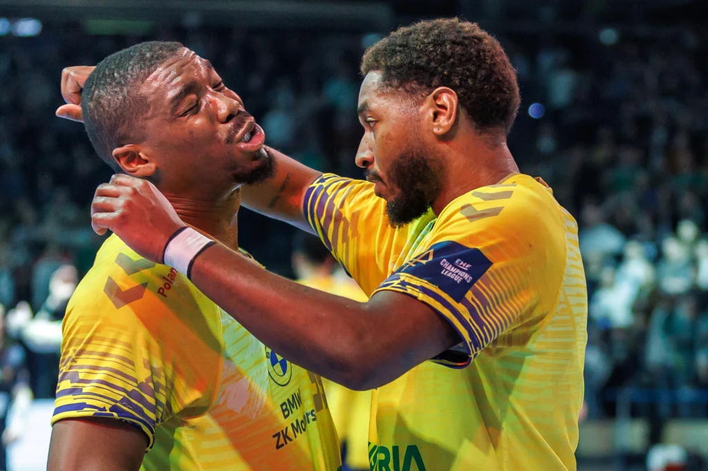 Szalona końcówka meczu Kielc w Lidze Mistrzów Dwóch szczypiornistów w żółtych koszulkach celebruje emocjonalny moment, obejmując się na boisku podczas meczu, wyraźnie odczuwając silne emocje. Tło stanowi niewyraźna publiczność na trybunach.