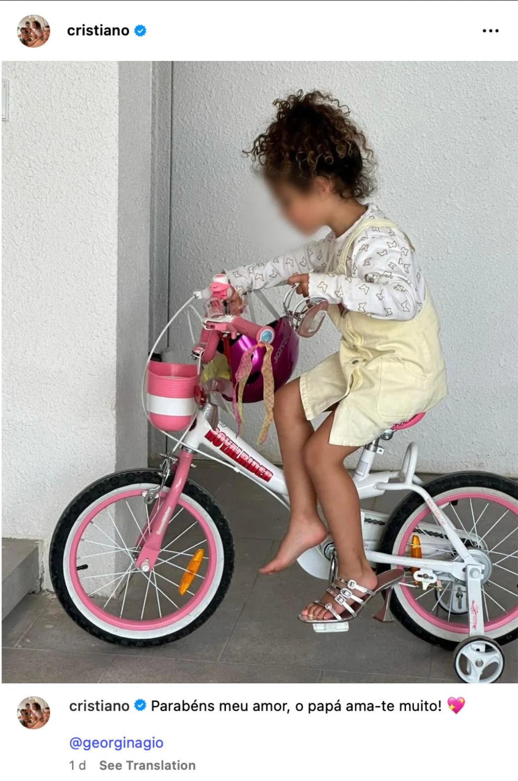 Dziewczynka z kręconymi włosami siedzi na różowym rowerku dziecięcym, ubrana w jasną sukienkę i sandałki, w tle jasna ściana.