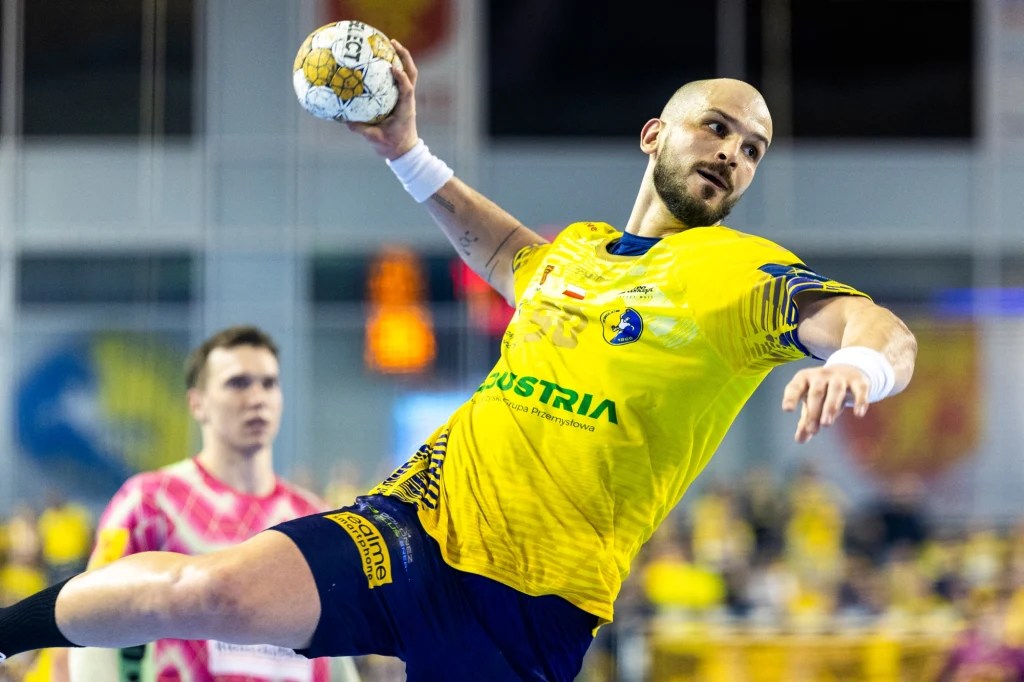 Zawodnik w żółtej koszulce z piłką w ręce wykonuje dynamiczny rzut podczas meczu piłki ręcznej, w tle widoczny jest inny zawodnik oraz publika.