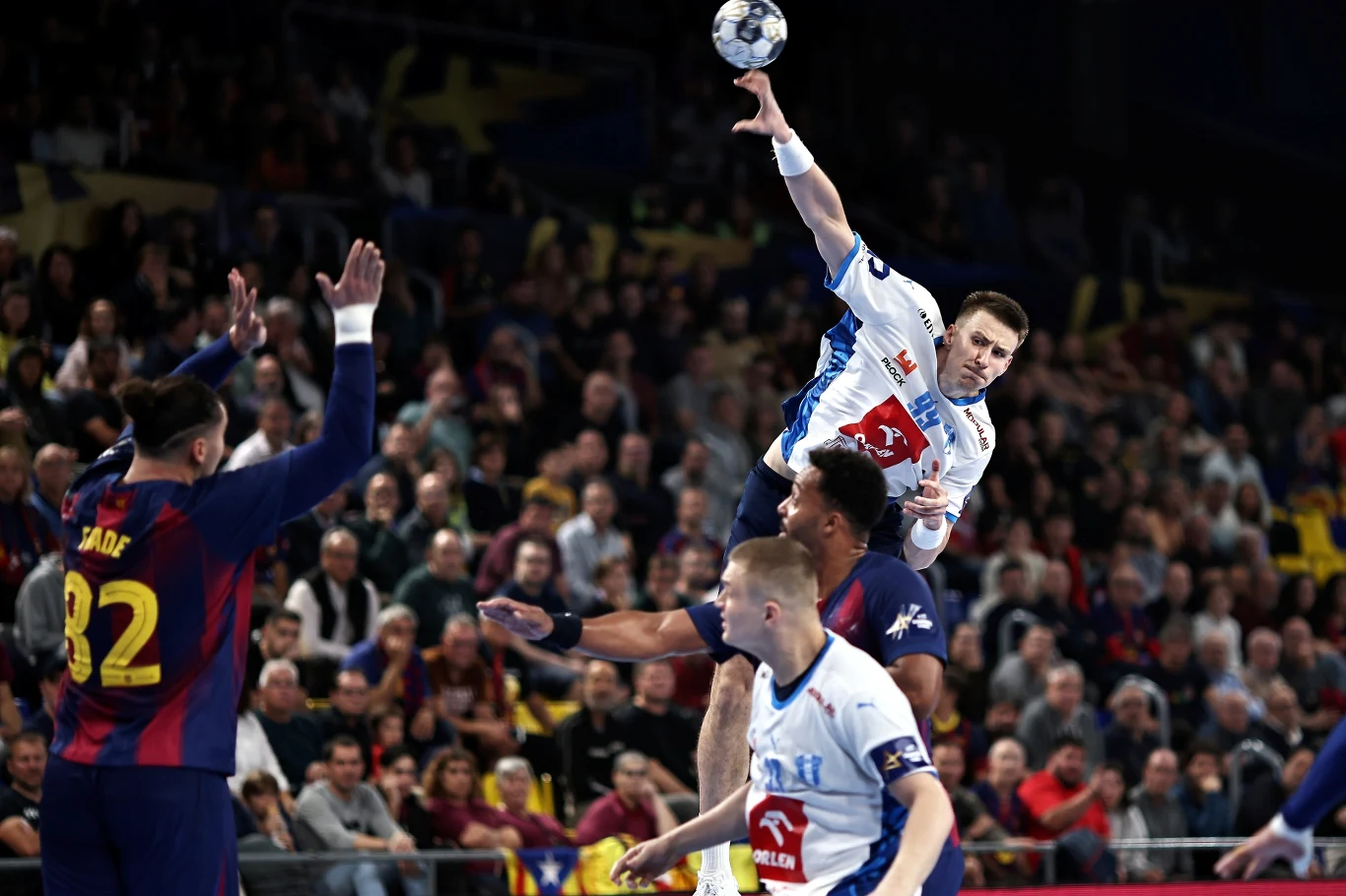 Zawodnik w białej koszulce wykonuje dynamiczny rzut piłki ręcznej w kierunku bramki, otoczony przez obrońców w granatowo-czerwonych strojach, tłum kibiców obserwuje grę z trybun.