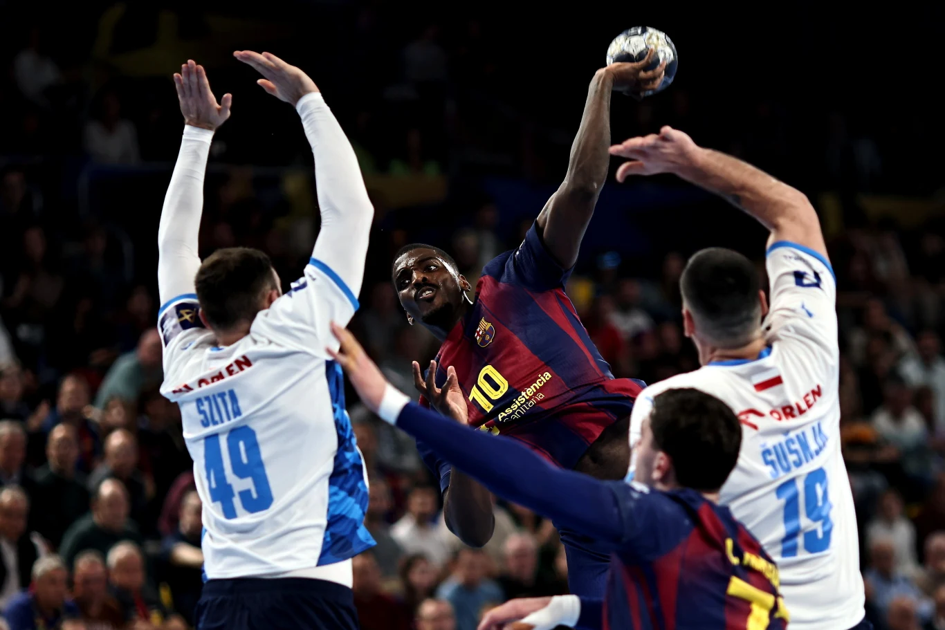 Intensywna akcja w meczu piłki ręcznej, zawodnik drużyny FC Barcelona z piłką w ręku usiłuje rzucić na bramkę, będąc otoczonym przez trzech zawodników drużyny przeciwnej w białych strojach. Na trybunach widoczna licznie zgromadzona publiczność.