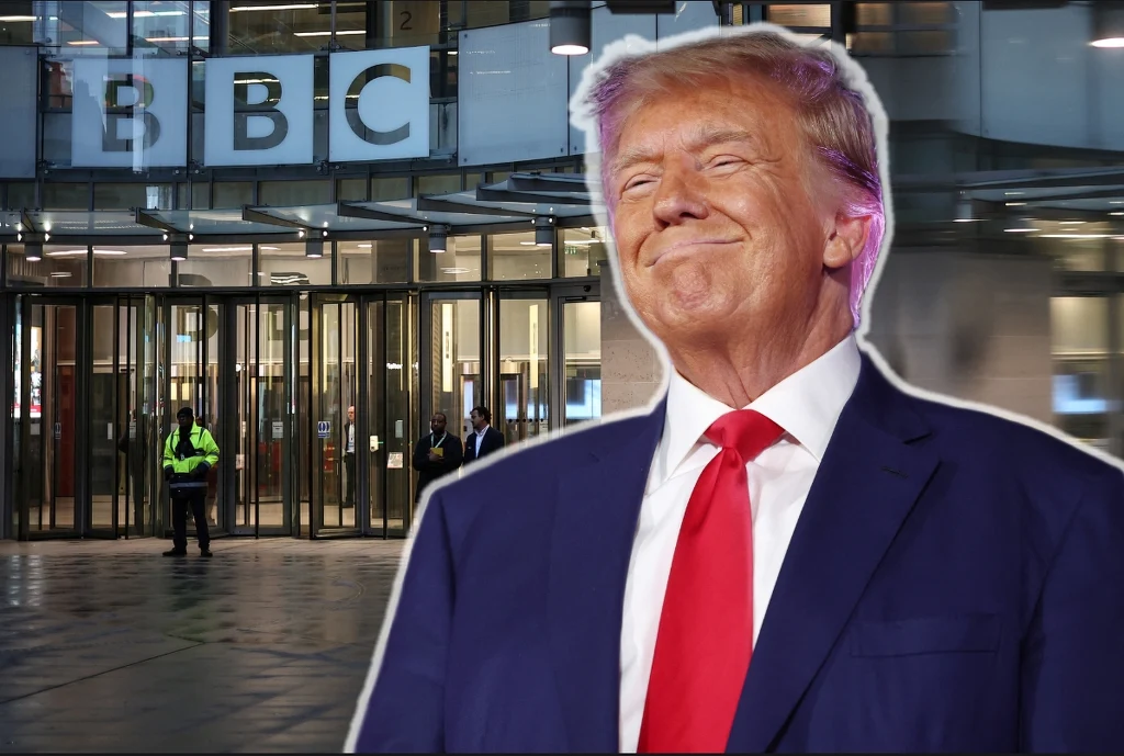 Uśmiechnięty mężczyzna w garniturze z czerwoną krawatą stoi na tle szklanych drzwi wejściowych do budynku BBC, w tle widoczny ochroniarz i logo stacji.