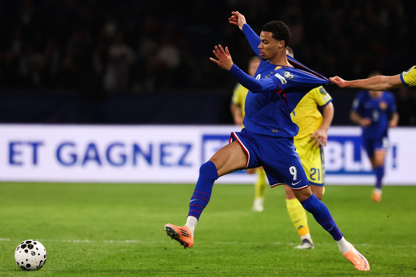 Piłkarz w niebieskim stroju z numerem 9 próbuje utrzymać się przy piłce, podczas gdy rywal w żółtej koszulce chwyta go za koszulkę, wyraźnie hamując jego ruch. Akcja dzieje się na tle zielonego boiska i widocznej publiczności.