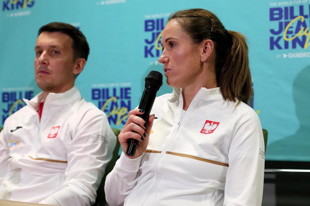 Dwoje sportowców w białych dresach z orłem na piersi siedzi przed niebieskim tłem z logo Billie Jean King Cup, kobieta trzyma mikrofon i udziela wypowiedzi.