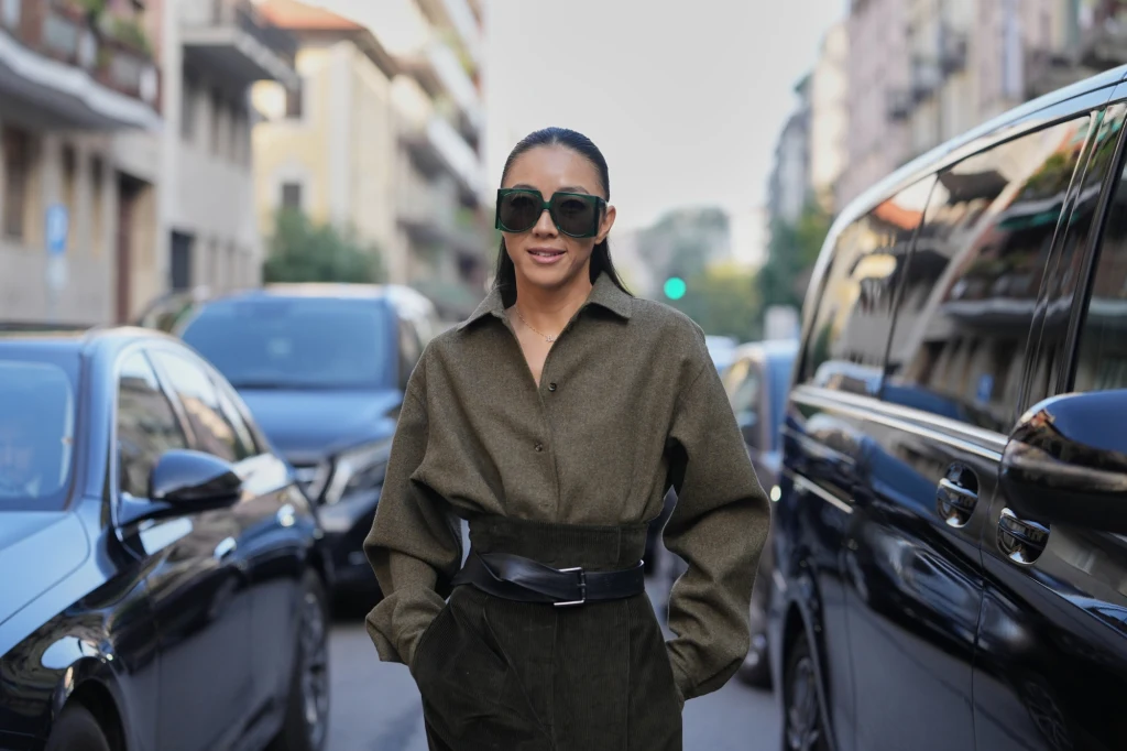 Kobieta w szerokich okularach przeciwsłonecznych stojąca na ulicy pomiędzy samochodami, ubrana w elegancką, oliwkową koszulę i ciemne spodnie z wysokim stanem, tło stanowią budynki mieszkalne i zaparkowane pojazdy.