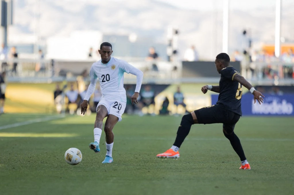 Dwóch piłkarzy w trakcie meczu rywalizuje o piłkę na boisku, jeden z zawodników ubrany na biało prowadzi piłkę, a przeciwnik w czarnym stroju zbliża się do niego, próbując ją przejąć. W tle widoczne są trybuny i rozmyte sylwetki widzów.