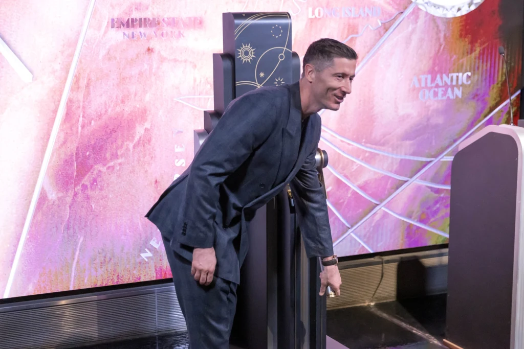 Elegancko ubrany mężczyzna w granatowym garniturze stoi pochylony przy instalacji na tle barwnej ściany z mapą i napisami. Na pierwszym planie dominują elementy scenografii, w tym geometryczna konstrukcja oraz fragmenty paneli.