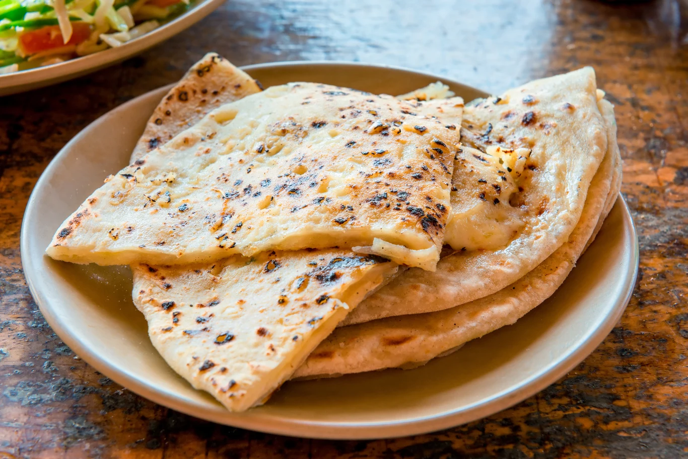 Chleb naan z masłem i czosnkiem to niekwestionowany ulubieniec świata Kilka kawałków ciepłego indyjskiego chlebka naan ułożonych na talerzu, na stole widoczna częściowo miska z sałatką warzywną.