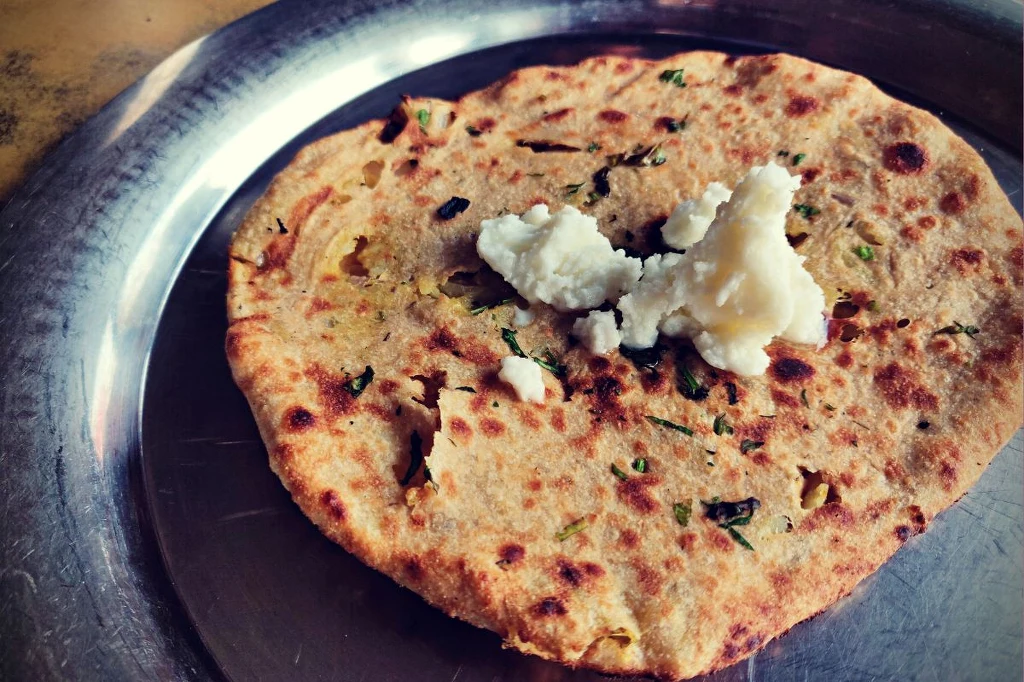 Kulcha z Pendżabu zajęła drugie miejsce w rankingu Taste Atlas Indyjski placek paratha z ziołami i nadzieniem, podany na metalowym talerzu z porcją masła na wierzchu.