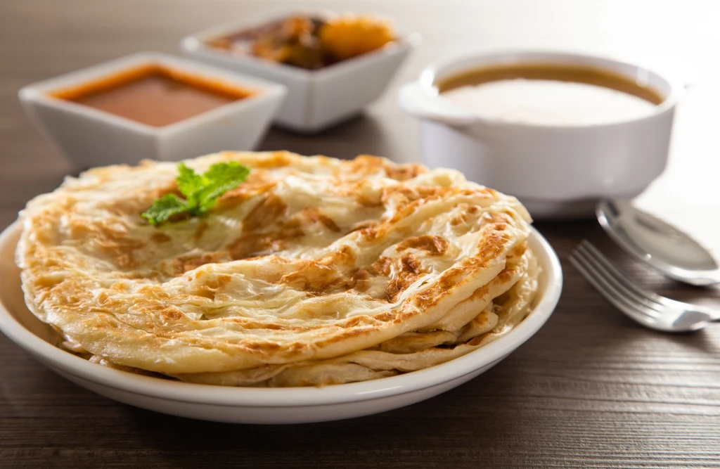 Roti canai zamyka pierwszą trójkę w rankingu Taste Atlas Stos placków roti canai na białym talerzu, udekorowany listkiem świeżej pietruszki, w tle trzy miseczki z różnymi sosami i dodatkami, obok widelczyk, całość ustawiona na drewnianym stole.
