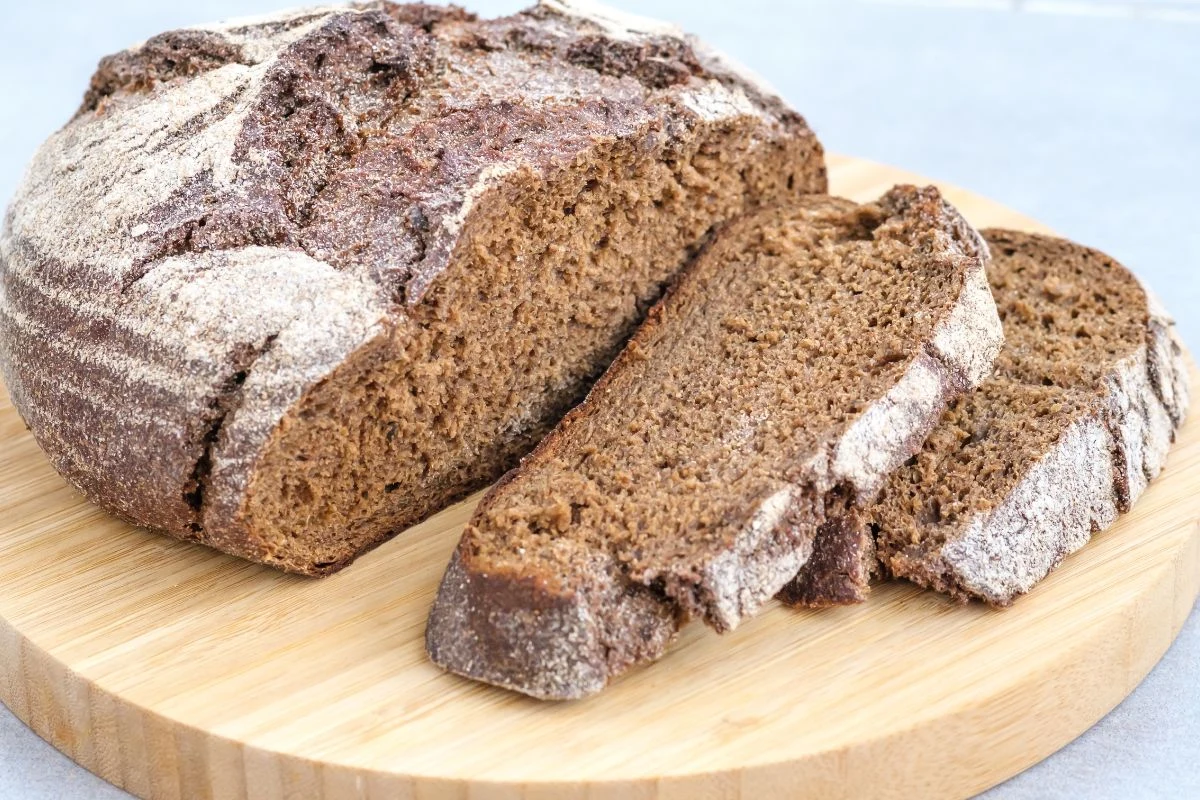 Niestety nasze cudowne żytnie bochny uplasowały się niemal na końcu listy Taste Atlas Bochenek domowego chleba razowego częściowo pokrojony na jasnobrązowej drewnianej desce. Chleb ma chrupiącą, lekko popękaną skórkę i ciemniejsze wnętrze.