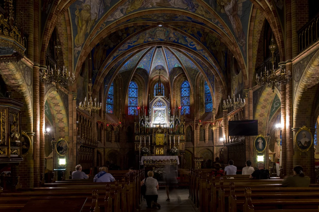 Kościół wchodzi w nowy okres/ zdj. ilustracyjne Wnętrze zabytkowego kościoła z wysokim sklepieniem, ozdobionymi freskami oraz kolorowymi witrażami. W centrum ołtarz główny podświetlony delikatnym światłem, wokół którego modli się kilka osób na ławkach i na kolanach. Atmosfera miejsca sakralnego podk...