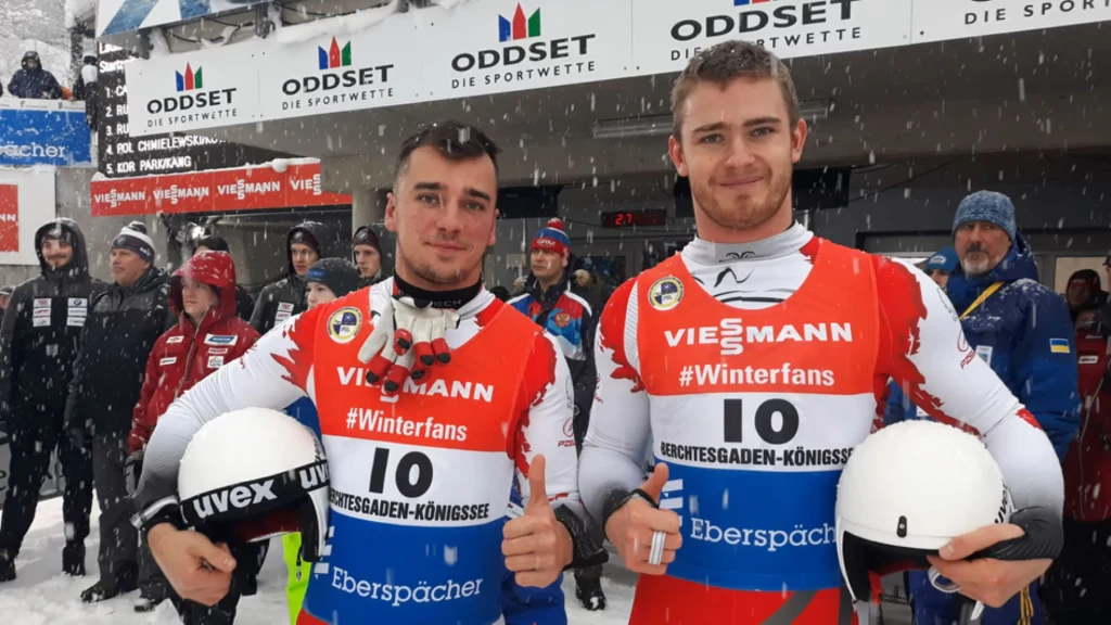 Dwóch sportowców w zimowych kombinezonach sportowych trzyma kaski narciarskie i pozuje do zdjęcia na tle zaśnieżonego stadionu, obok widoczni kibice i ekipy techniczne.