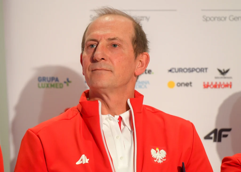 Mężczyzna w czerwonej kurtce z białym orłem oraz logo marki 4F na piersi, siedzący przed tablicą z logotypami sponsorów i partnerów.