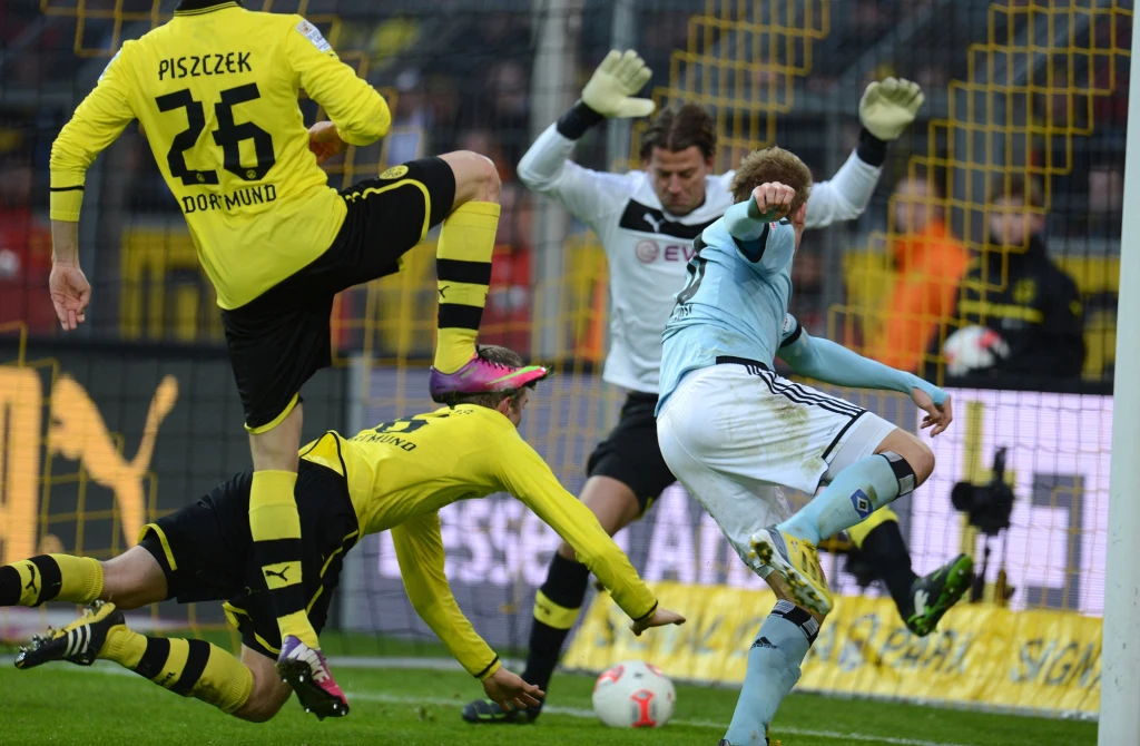 Artjoms Rudnevs w Bundeslidze mierzył się m.in. z Borussią Dortmund z Jakubem Błaszczykowskim, Robertem Lewandowski i Łukaszem Piszczkiem w składzie Dwóch piłkarzy w żółto-czarnych strojach próbuje zablokować zawodnika w błękitnej koszulce, który oddaje strzał na bramkę, bramkarz w białej bluzie jest gotowy do interwencji na tle siatki bramkowej.