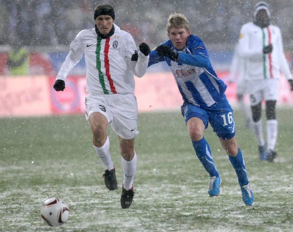 Artjoms Rudnevs w barwach Lecha Poznań toczył niezapomniane boje w Lidze Europy Dwóch piłkarzy biegnie za piłką po śnieżnym boisku, ubrani w stroje odpowiednich drużyn, w tle widoczny intensywny opad śniegu i zamazane sylwetki innych zawodników.