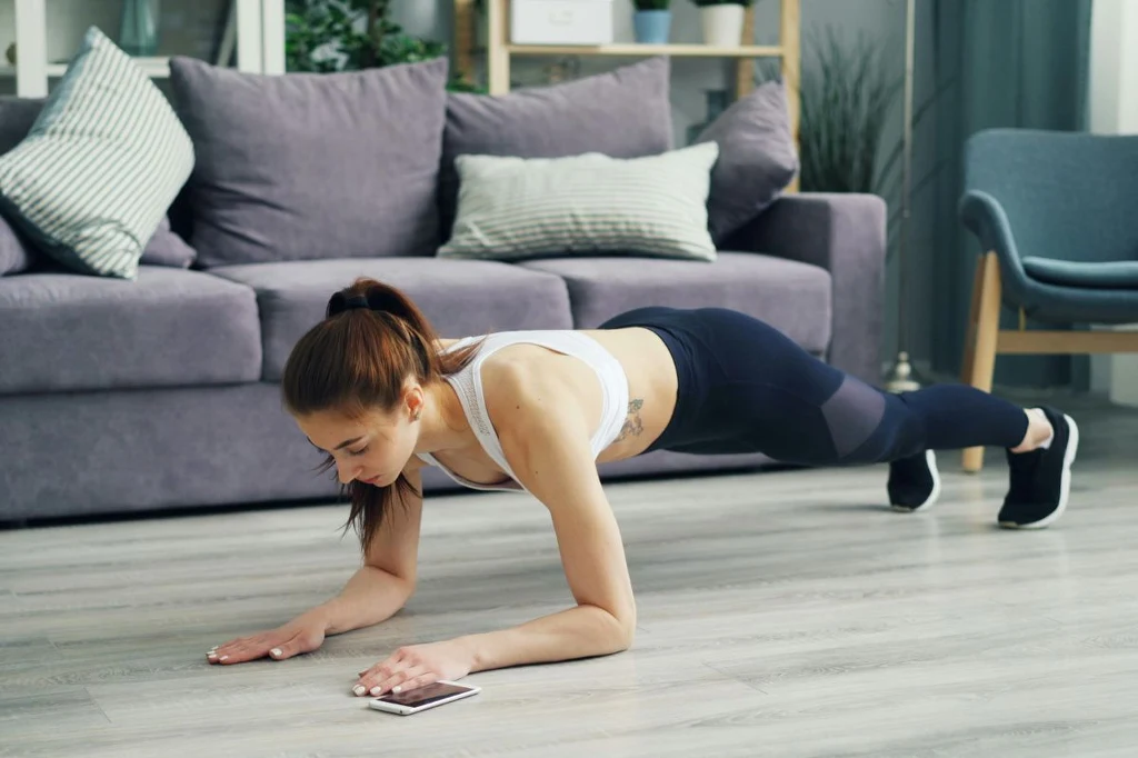 Plank to świetne ćwiczenie na mięśnie głębokie. Możesz robić je w domu Kobieta w sportowym stroju wykonuje ćwiczenie typu deska w salonie, opiera się na przedramionach i palcach stóp, obok leży smartfon, w tle sofa i rośliny.