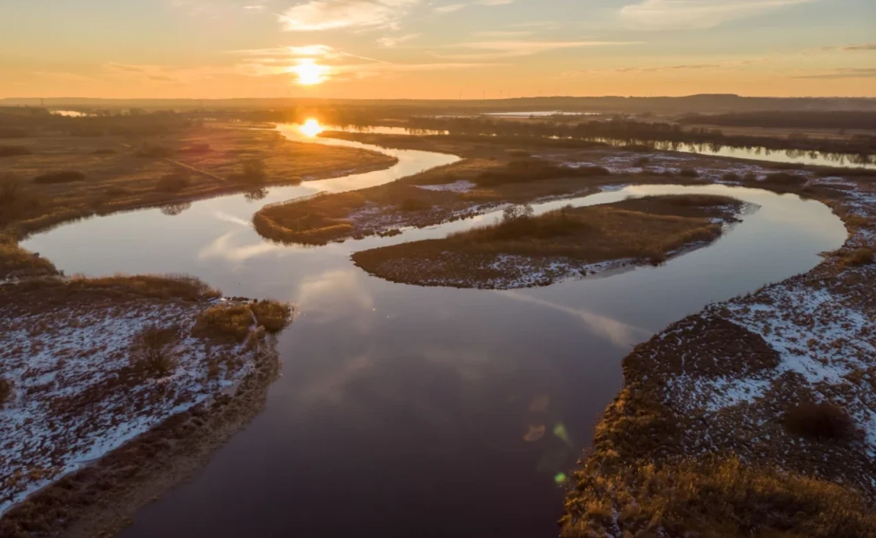 W Międzyodrzu występuje 237 gatunków ptaków, co stanowi 50 proc. wszystkich gatunków odnotowanych w Polsce/ fot. Piotr Piznal / Ministerstwo Klimatu i Środowiska Kręta rzeka przepływająca przez szerokie, częściowo pokryte śniegiem łąki, oświetlona promieniami zachodzącego słońca. Na horyzoncie rozciągają się delikatne pasma wzgórz, a złote światło odbija się w wodzie.