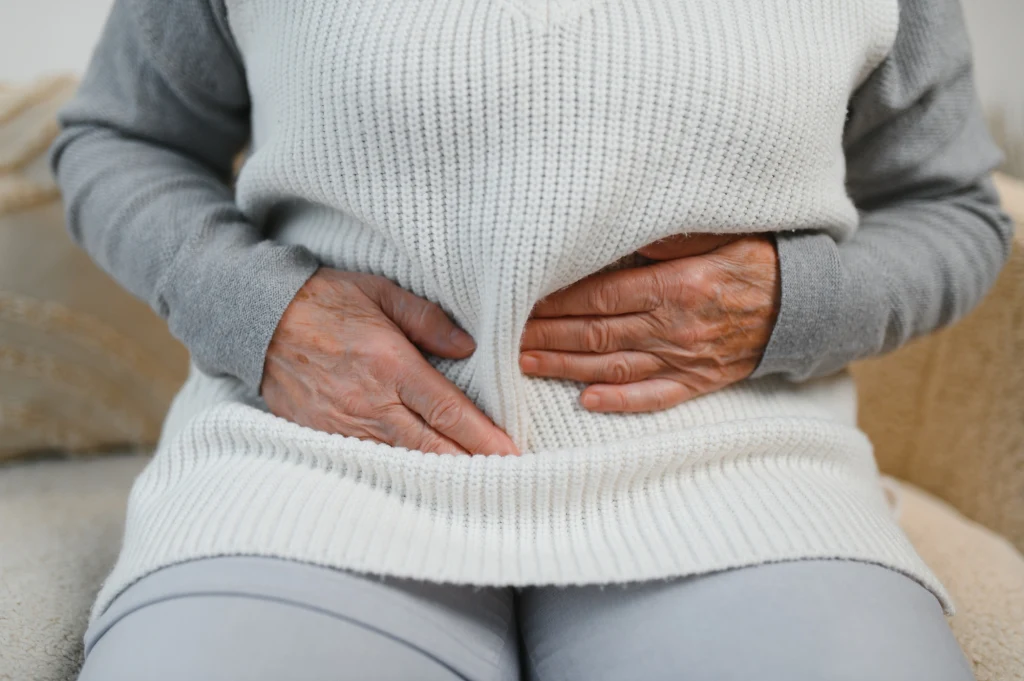 Mięśnie głębokie odpowiadają m.in. za kontrolę nad pęcherzem Starsza osoba trzymająca się oburącz za brzuch, ubrana w jasny sweter i spodnie, wyrażająca dyskomfort lub ból.