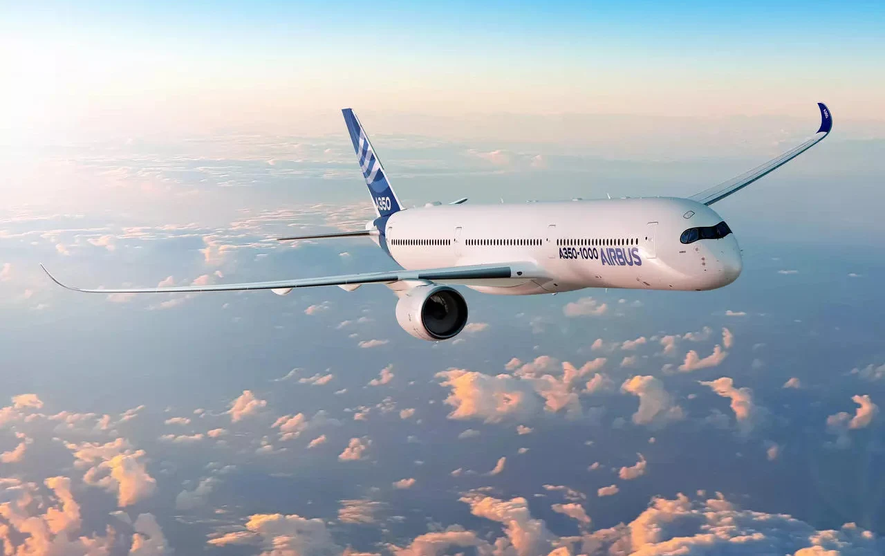 Najdłuższy lot samolotem. W trakcie podróży dwa wschody Słońca. Nowoczesny samolot pasażerski Airbus A350-900 leci wysoko ponad chmurami w trakcie dnia, skrzydła rozpostarte szeroko, niebo jest jasne, z lekkim zabarwieniem od zachodzącego słońca, a pod nim rozciąga się panoramę chmur.